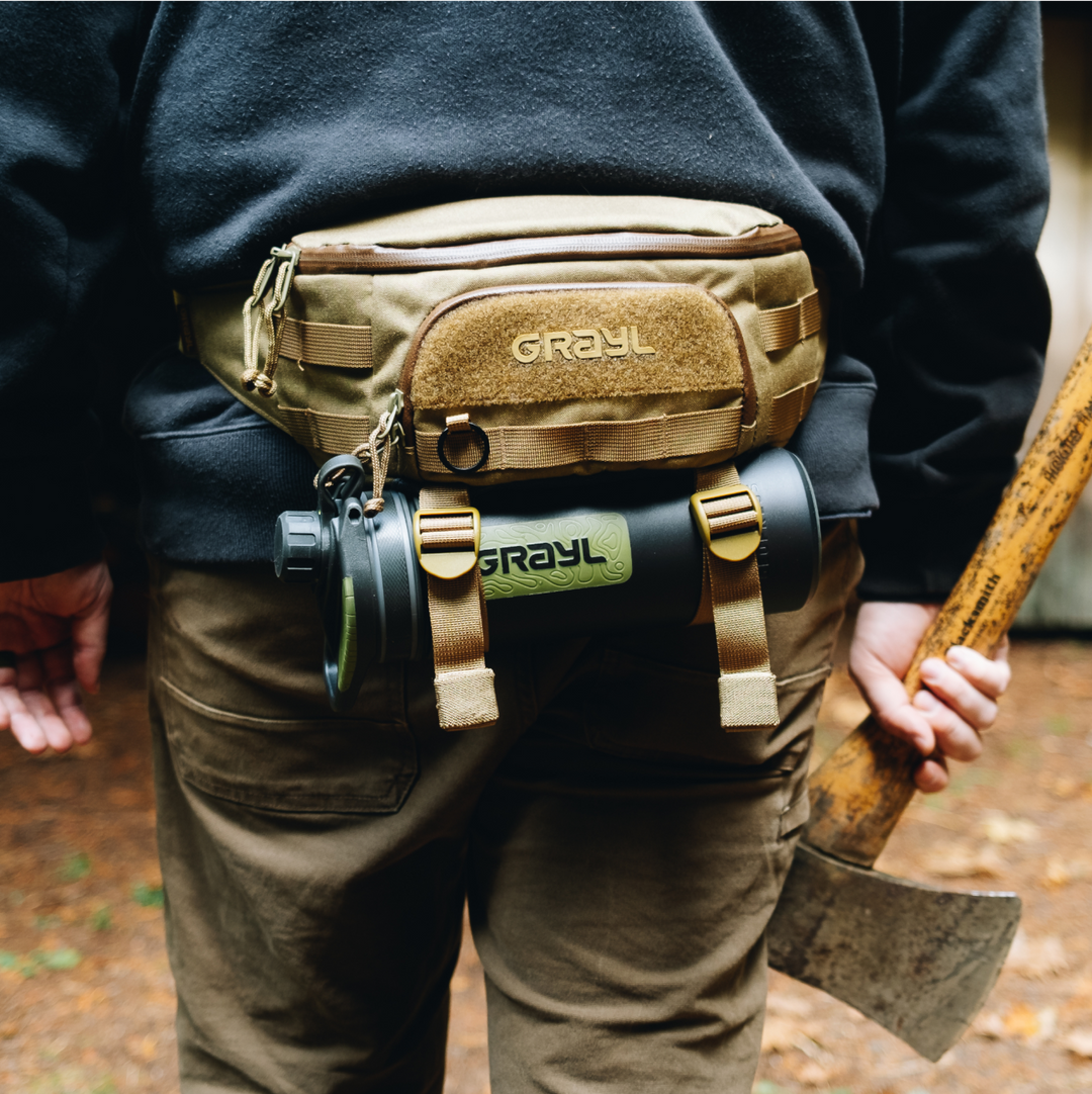 2.5L Transport Hip Pack in Coyote Brown worn around the waste of someone camping and holding an ae in had. GeoPress Black Camo water bottle suspended in BottleLock Tie Down System.