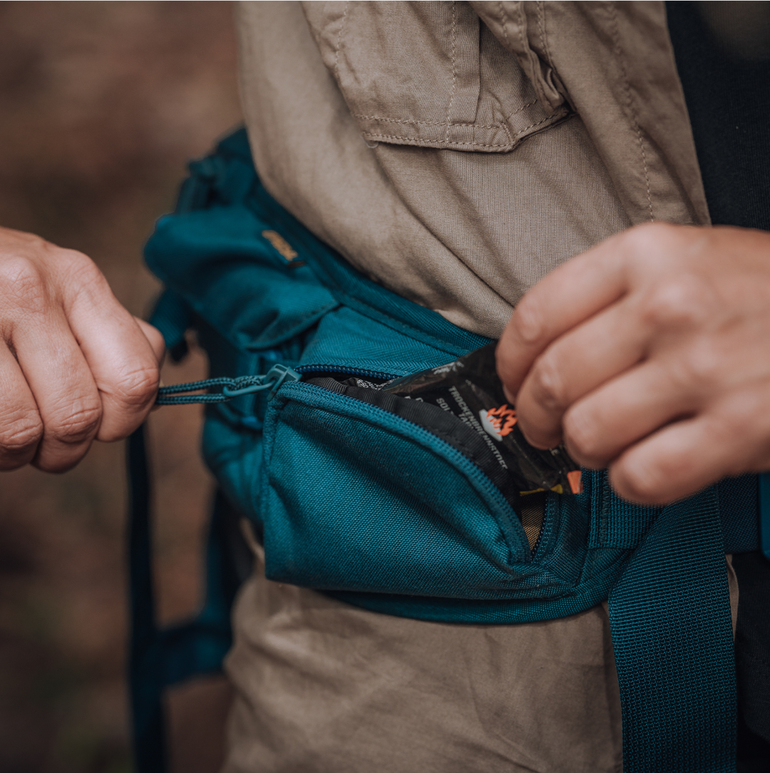 Male hiker wearing hiking clothing and a Grayl Mission Hip Pack in Wander Blue colorway. Demonstrating front zipper pocket functionality.