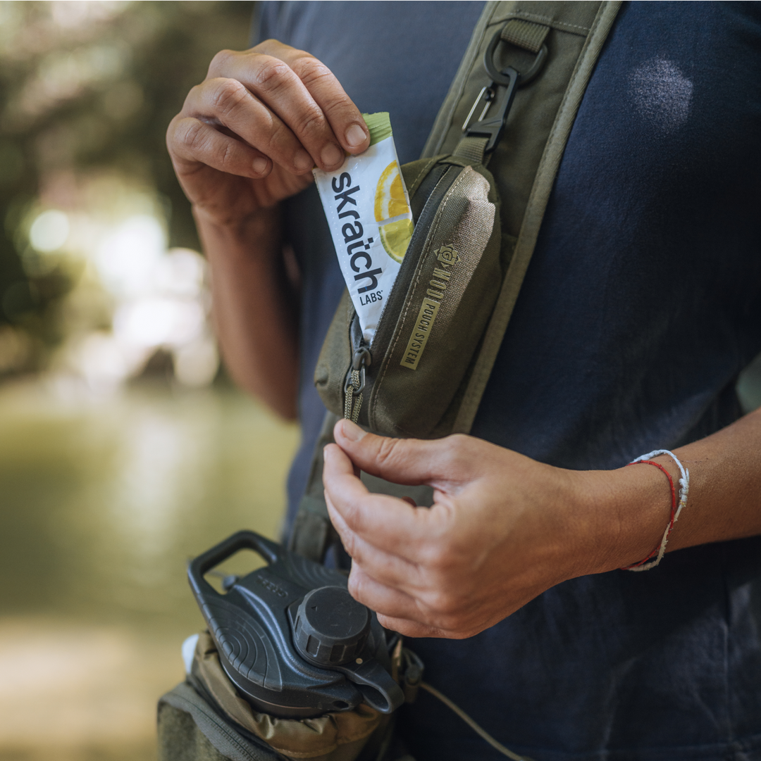 Mission Bottle Carrier and G-MOD Pod Pouch in Olive Drab on the front-side of a Grayl adventurous hiker. GeoPress Titanium filter and purifier bottle visible in main compartment. G-MOD Pod Pouch is zipped open with Skratch Labs drink-mix single being pulled out.