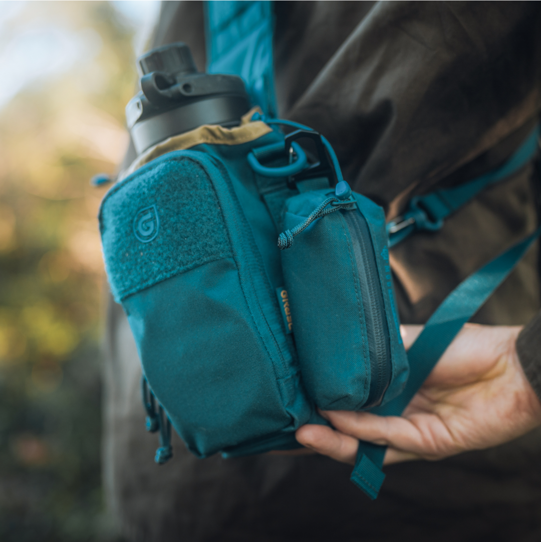 Transport Bottle Carrier and G-MOD Pod Pouch in Wander Blue on the front-side of a Grayl adventurous hiker. GeoPress Titanium filter and purifier bottle visible in main compartment.