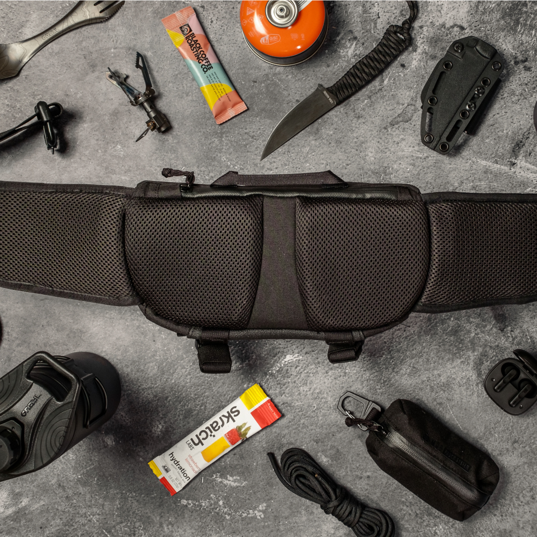 Flat lay of Grayl Mission Hip Pack in Covert Black colorways. Knife, sheath, fuel canister, Black Coffee Roasting Company single serving, Skratch Labs electrolyte drink mix, Grayl GeoPress, Grayl Titanium Stove and Ti Spork visible.