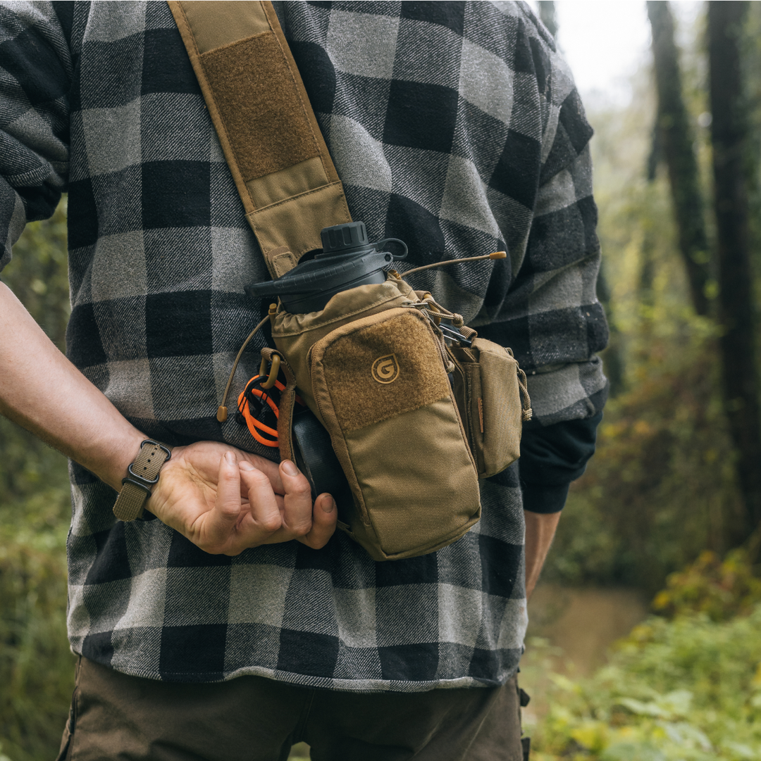 Hiker wearing Grayl® Transport Bottle Sling in Coyote Brown colorway. Male hiker is accessing cell phone from full-zip pocket while on trail. GeoPress water filter purifier bottle visible in main compartment.